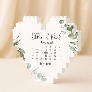 May include: A white heart-shaped block calendar with the names "Ellie & Paul" and the word "Engaged" printed on it. The calendar highlights July 2025, with the 15th circled. Greenery accents the design.