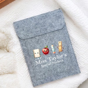 May include: Grey felt laptop sleeve featuring a white graphic of a smiling apple, pencil, ruler, and notebook. The text "Miss Taylor's Special Stickers" is printed below the graphic.