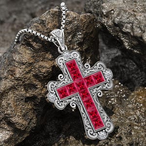 May include: A silver pendant necklace featuring a cross design. The cross is adorned with red gemstones and surrounded by an ornate, filigree-style border. Small, clear gemstones are set around the edges. The necklace rests on a textured, dark gray rock surface.