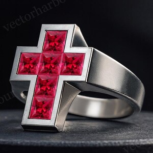May include: A silver ring featuring a cross design. The cross is filled with square, bright red gemstones. The ring is set against a dark background, highlighting the contrast between the silver and the red stones.