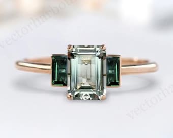 May include: A rose gold engagement ring featuring a large emerald cut centre stone and two smaller emerald cut green stones on either side.