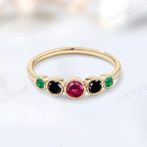 Puede incluir: Un anillo de oro con una piedra preciosa roja central, flanqueada por dos piedras preciosas negras y dos piedras preciosas verdes. El anillo tiene una banda delgada y un acabado pulido, que resalta los colores vibrantes de las piedras preciosas.