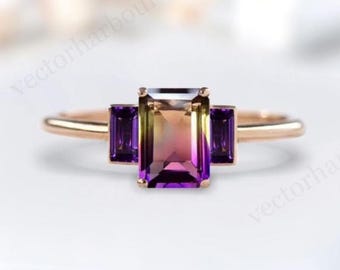 May include: A rose gold ring featuring a rectangular, multi-colored gemstone with purple and yellow hues, flanked by two smaller, rectangular purple gemstones. The ring is set against a soft, blurred background.