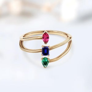 Może przedstawiać: Złoty pierścionek z trzema kolorowymi kamieniami szlachetnymi: różowym kamieniem w kształcie markizy, prostokątnym niebieskim kamieniem i zielonym kamieniem w kształcie gruszki. Pierścionek ma podwójną opaskę.