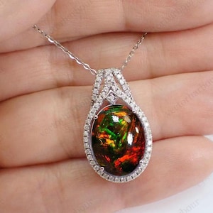 May include: A teardrop-shaped pendant necklace featuring a large, oval-cut black opal with vibrant red and green flashes. The stone is surrounded by a halo of small, clear pavé-set diamonds, set in a polished silver-toned metal frame on a chain.