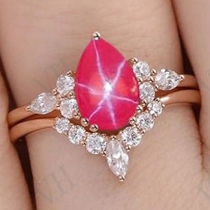 Könnte beinhalten: Ein roségoldener Verlobungsring mit einem roten Sternrubin in Tropfenform und einem Heiligenschein aus kleineren Diamanten. Der Ring hat eine passende Trauring mit einer Reihe von Diamanten.