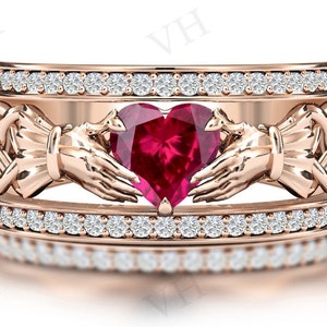 Pode incluir: Um anel de ouro rosa com uma grande gema vermelha em forma de coração cercada por duas mãos que seguram o coração. O anel é adornado com uma fileira de pequenas gemas transparentes de cada lado do coração.