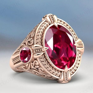 以下が含まれることがあります： 複雑な花柄で囲まれた大きな楕円形のルビーをセットしたゴールドリング。リングの側面には小さなルビーのアクセントが施されています。