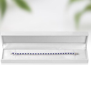 May include: A white jewellery box containing a blue sapphire tennis bracelet. The bracelet is made of silver and has a clasp.