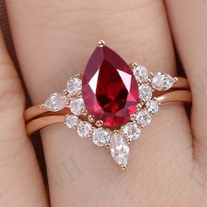 May include: A pear-shaped ruby engagement ring set in rose gold with a halo of smaller diamonds. The ring is accented with additional pear-shaped diamonds on the band.