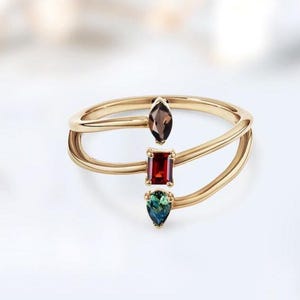 May include: A gold ring with three gemstones: a brown marquise-cut stone, a red rectangular stone, and a teal pear-shaped stone. The ring has a unique double-band design, with the gemstones set in the center.