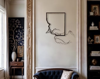 Op de afbeelding: Een vierkant wandkunstwerk met een zwart frame en een takvormig ontwerp in het frame. De kunst hangt aan een witte muur in een kamer met een donkerblauwe fauteuil en een houten boekenkast.