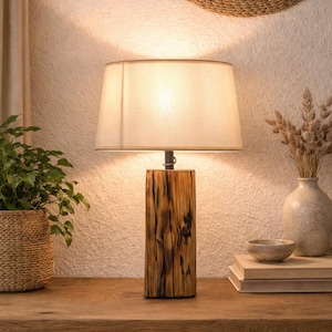 Op de afbeelding: Tafellamp met een beige lampenkap en een voet van donkerbruin, getextureerd hout. De lamp staat op een houten tafel, samen met een plant in een gevlochten mand en decoratieve items.