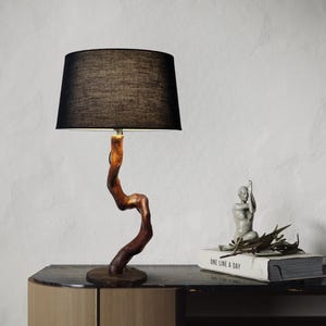 May include: A table lamp with a black fabric shade and a wooden base shaped like a twisted branch. The lamp is on a wooden table with a book and a small statue on top.