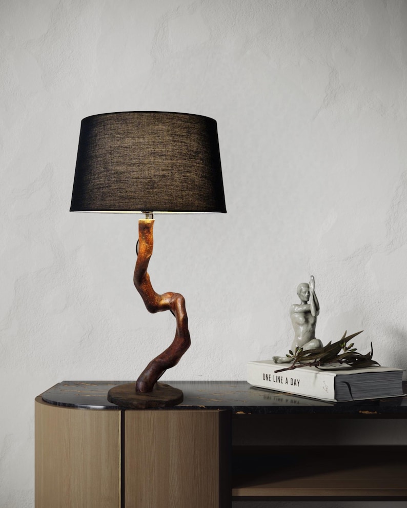 May include: A table lamp with a black fabric shade and a wooden base shaped like a twisted branch. The lamp is on a wooden table with a book and a small statue on top.