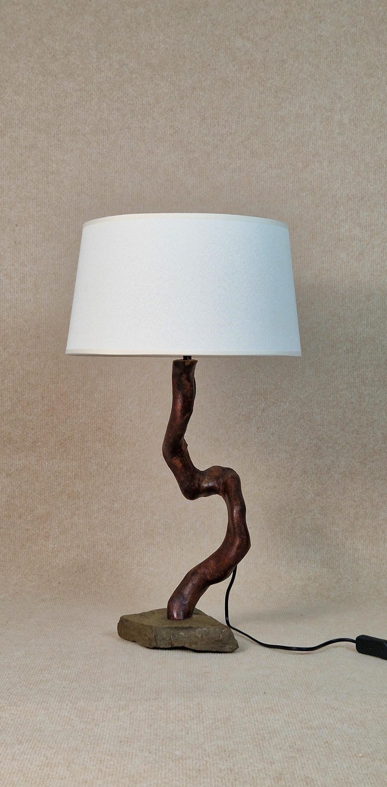 May include: A table lamp with a white fabric lampshade and a wooden base shaped like a twisted branch. The base is mounted on a flat piece of stone.