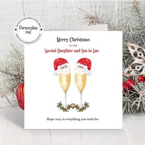 May include: A square Christmas greeting card featuring two champagne flutes, each topped with a red Santa hat labeled Sarah and John. The text reads Merry Christmas to our Special Daughter and Son in Law and Hope 2027 is everything you wish for.
