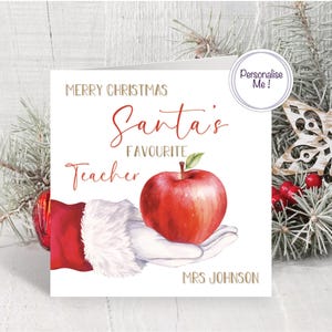 May include: A Christmas card with the text "Merry Christmas Santa's Favourite Teacher". The card features a red apple held in a Santa Claus hand. The card is personalized with the name "MRS JOHNSON".