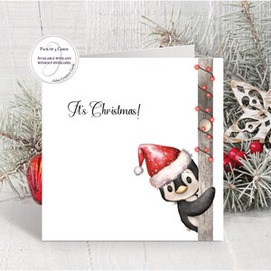 May include: A square greeting card featuring a watercolor illustration of a penguin wearing a red and white Santa hat, peeking from behind a wooden post wrapped in red string lights. The text It's Christmas! is printed in black script.