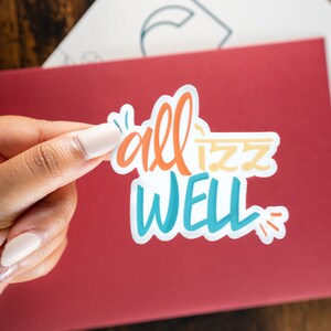 May include: A white sticker with the phrase "all is well" written in colourful, stylized lettering. The sticker is being held by a hand.