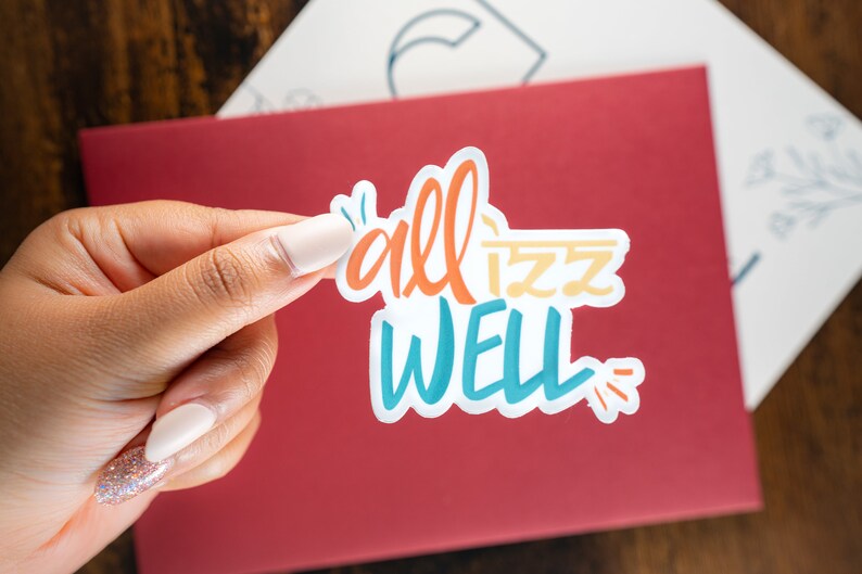 May include: A white sticker with the phrase "all is well" written in colourful, stylized lettering. The sticker is being held by a hand.