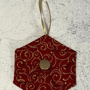 Könnte beinhalten: Ein roter, sechseckiger Weihnachtsbaumschmuck aus Stoff mit goldenem Wirbelmuster. Er hat einen goldenen Knopf in der Mitte und ein goldenes Band zum Aufhängen. Eine festliche Dekoration für die Weihnachtszeit.