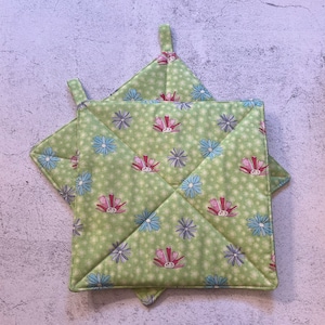May include: Two square, quilted pot holders with a light green background and a floral pattern. The flowers are pink, blue, and purple. Each pot holder has a small loop for hanging.