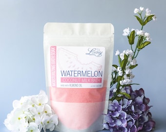 May include: A white pouch of watermelon coconut milk bath soak with a pink and white label. The label features a watermelon slice graphic. The pouch is surrounded by white and purple flowers against a light blue background.