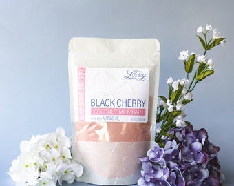 May include: A white pouch with a pink label that reads "Black Cherry Coconut Milk Bath" with the text "Made with Almond Oil" and "e 8.8 oz (250 g)". The pouch is filled with a pink bath soak and is surrounded by white and purple flowers.