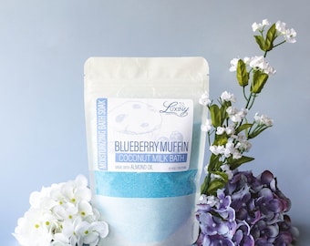 May include: A white pouch with a blue bath soak inside. The pouch has a label that says "Blueberry Muffin Coconut Milk Bath" and "Made with Almond Oil". The pouch is surrounded by white and purple flowers.