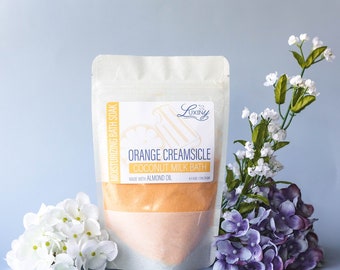 May include: A white pouch with a label that reads "Orange Creamsicle Coconut Milk Bath" and "Made with Almond Oil". The pouch contains a bright orange bath soak. The pouch is sitting on a light blue surface with white and purple flowers.