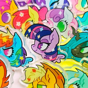 Op de afbeelding: Een verzameling kleurrijke cartoon pony-personage pins. De pins zijn gemaakt van helder acryl met een bedrukte afbeelding van een pony. De pony's zijn allemaal in verschillende kleuren en hebben verschillende uitdrukkingen.