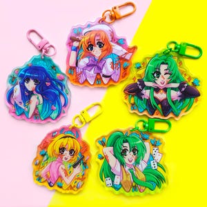 May include: Four colorful anime character keychains with different designs. Each keychain features a female character with a unique outfit and expression. The keychains are made of clear acrylic and have a colorful background with butterflies and other details.