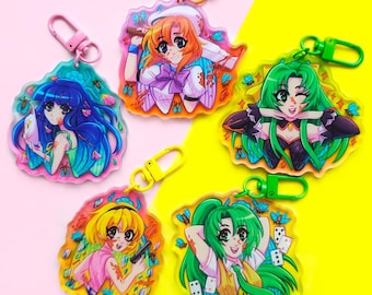 May include: Four colorful anime character keychains with different designs. Each keychain features a female character with a unique outfit and expression. The keychains are made of clear acrylic and have a colorful background with butterflies and other details.