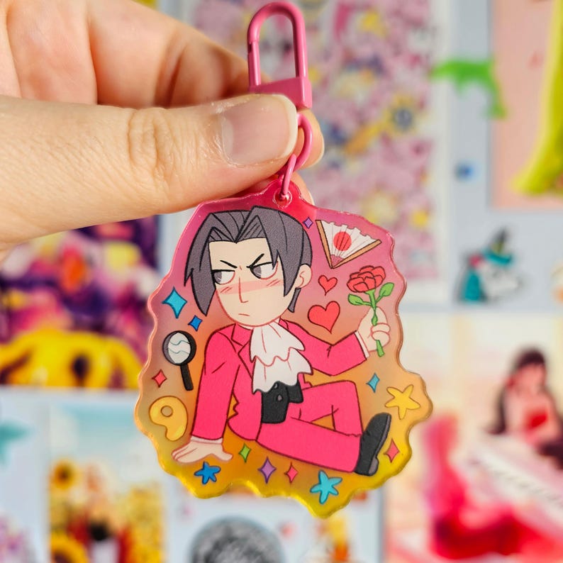 May include: A pink and yellow anime-style keychain featuring a character with black hair, a pink suit, and a rose. The keychain has a pink metal clasp.