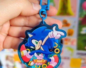 May include: A blue and red cartoon character keychain featuring Sonic the Hedgehog running with a golden ring and a hot dog. The keychain is made of clear acrylic and has a blue metal ring.