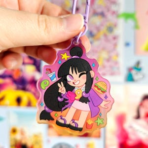 May include: A pink and purple character charm with a cartoon girl wearing a purple jacket and a pink bow. The charm has a glittery background and features a drink, a burger, and fries.