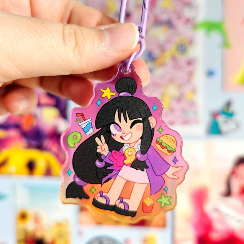 May include: A pink and purple character charm with a cartoon girl wearing a purple jacket and a pink bow. The charm has a glittery background and features a drink, a burger, and fries.