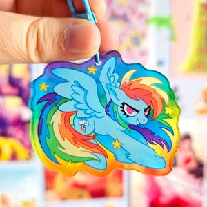 May include: A rainbow-coloured plastic charm featuring a cartoon image of Rainbow Dash, a winged pony from the My Little Pony franchise, with a lightning bolt symbol on her flank. The charm is shaped like a cloud and has a holographic finish.