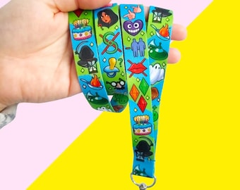 May include: A blue and green lanyard with a variety of colorful cartoon images, including a birthday cake, a ghost, a heart, a witch, and a pair of glasses. The lanyard has a metal clip at the end.