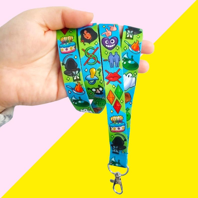May include: A blue and green lanyard with a variety of colorful cartoon images, including a birthday cake, a ghost, a heart, a witch, and a pair of glasses. The lanyard has a metal clip at the end.