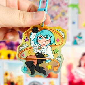 May include: A holographic charm featuring a cartoon character with blue hair, a white shirt, and black pants. The character is holding a yellow rope and is surrounded by stars and other colorful shapes.