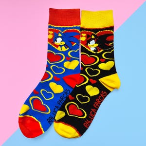 May include: A pair of colorful socks featuring cartoon characters and heart designs. One sock is blue with red and yellow accents, the other is black with yellow and red accents. The text "RALUCA FARCAS" is printed on each sock.