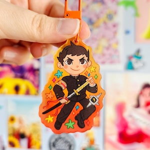 May include: A colorful anime-style character keychain with a sword and a star-shaped background. The character is wearing a black uniform with a red stripe on the sleeve. The keychain is made of clear acrylic and has a shiny finish.