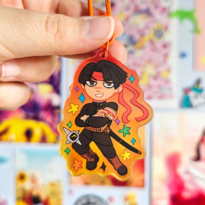 May include: A colorful anime-style character keychain with a red headband and a sword. The character is wearing a black uniform and is standing on a yellow background with stars.