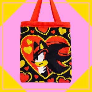 May include: A knitted tote bag with a black, red, and yellow design. The bag features a heart-shaped graphic with a character's face. The bag has red handles and trim, set against a pink and yellow geometric background.