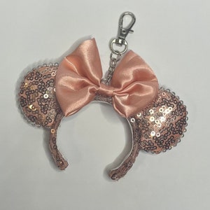 May include: A rose gold sequined ear headband with a matching satin bow, attached to a silver-tone keychain. The ears are covered in small, shiny sequins. The bow is centered on the headband, adding a touch of elegance.