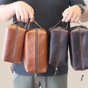 May include: Four leather zippered travel bags in brown, tan, and dark brown. The bags have a top handle and a zipper closure.