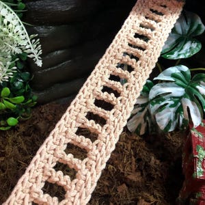 May include: A long, narrow strip of light beige crochet fabric featuring a repeating openwork pattern of rectangular gaps framed by thick, textured yarn borders. The piece is crafted with a consistent stitch structure throughout its entire length.