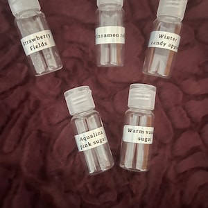 Puede incluir: Cinco botellas pequeñas de plástico transparente con etiquetas blancas. Las etiquetas dicen "Strawberry Fields", "Cinnamon Roll", "Winter candy apple", "Aqualina pink sugar" y "Warm van sugar". Las botellas tienen tapas abatibles blancas y están dispuestas sobre una tela burdeos oscura.